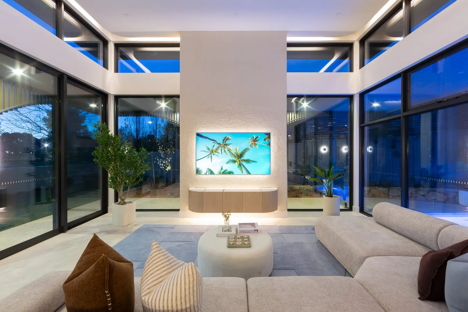 Open-plan living at dusk, a deep lounge under a high ceiling with glass on three sides