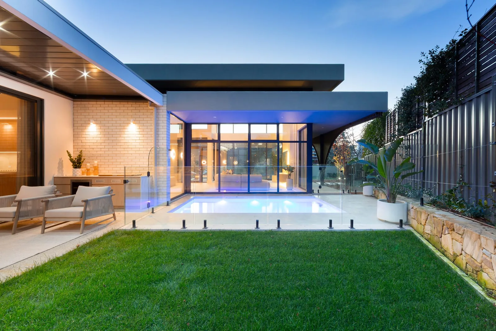 The heated pool at twilight, the lit house wrapped around the water and the lawn in front